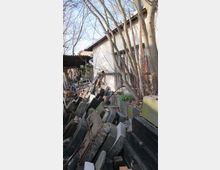 Ein Hofbereich mit gestapelten Grabsteinen und anderen Steinblöcken neben einem Gebäude mit hell verputzter Fassade und Fenstern. Im Vordergrund stehen Bäume ohne Laub, und im Hintergrund ist ein überdachter Bereich mit Gegenständen sichtbar.