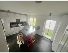 Moderne Küche mit weißen Einbauschränken, schwarzer Arbeitsplatte und zentralem Kochfeld auf einer Kücheninsel. Große Fenster und eine Glastür mit Sicht auf einen Garten lassen viel Tageslicht herein; der Boden ist mit dunklen Fliesen ausgelegt.