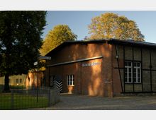 Backsteingebäude mit der Aufschrift „Exerzierhalle“, umgeben von Bäumen mit gelbem Laub. Im Vordergrund sind ein gepflasterter Weg, ein Zaun und ein Hinweisschild zu sehen.