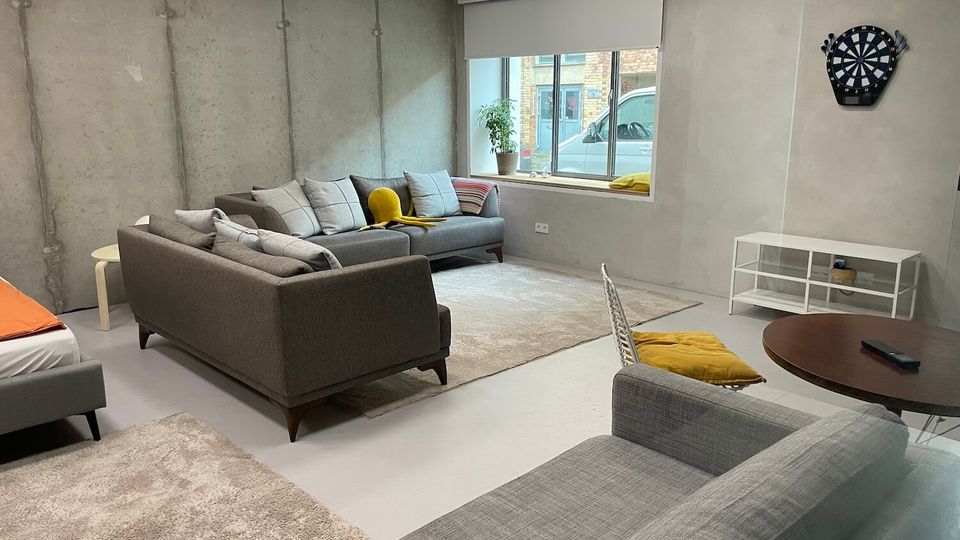 Industrial-style lounge room with exposed concrete walls, a large window with a roller blind, and gray sofas arranged on light rugs. A wall-mounted dartboard and a low white media unit sit against the far wall, with a small round table and chair in the foreground.