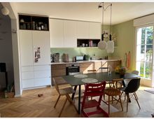 Moderne Küche mit Essbereich. Helle Fronten, Holzunterschränke und eine grüne Wand bilden die Küchenzeile, während ein großer Esstisch mit verschiedenen Stühlen im Vordergrund steht; rechts führt eine bodentiefe Glastür zu einem Garten.