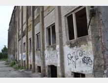 Fassade eines verlassenen Industriegebäudes aus weißem Ziegelstein, an dem mehrere Fenster beschädigt oder offen sind. Die Wände sind mit Graffiti bedeckt, und der Boden davor ist mit Betonplatten und etwas Vegetation bedeckt.