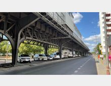 Ansicht einer erhöhten Stahlkonstruktion, die eine Bahnlinie trägt, unter der Autos und Wohnmobile geparkt sind. Im Hintergrund sind Bäume sowie ein Bürgersteig mit einer Bushaltestelle zu erkennen, daneben verläuft eine Straße.