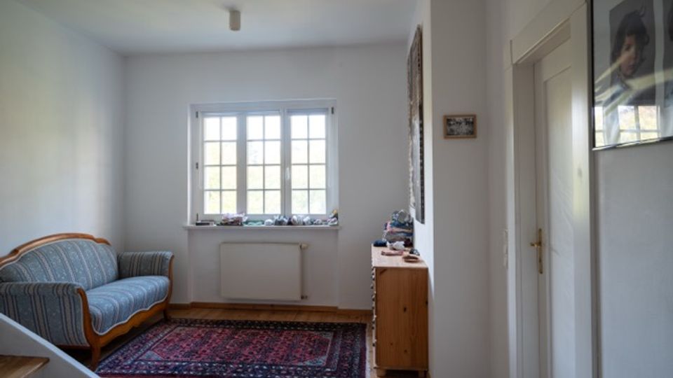 Schmaler, heller Wohnraum mit Holzfußboden, einem gemusterten Teppich und einem blau gestreiften Sofa an der linken Wand. Am Ende des Raums befindet sich ein mehrteiliges Fenster über einem Heizkörper; rechts stehen eine kleine Kommode und eine weiße Tür, an den Wänden hängen Bilder.