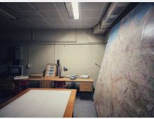 Kleiner, fensterloser Arbeitsraum mit niedriger Decke, Neonleuchte, sichtbaren Leitungen und einem langen Schreibtisch mit alten Kommunikationsgeräten und Telefon. Rechts lehnt eine große, detaillierte Landkarte an der Wand; vorne steht ein großer Arbeitstisch.