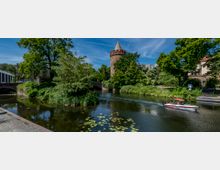 Flusslandschaft mit einem historischen Turm aus rotem Backstein, umgeben von üppigem Grün. Im Vordergrund fährt ein kleines Motorboot mit rotem Sonnendach, während Seerosen die Wasseroberfläche bedecken.