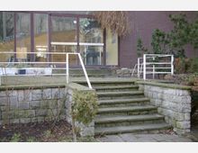 Außenansicht eines Gebäudeeingangs mit einer Steintreppe, die von niedrigen Steinmauern und einem weißen Metallgeländer flankiert wird. Im Hintergrund befindet sich eine große Glasfassade, die einen Blick ins Innere des Gebäudes ermöglicht, umgeben von vereinzeltem Pflanzenbewuchs.