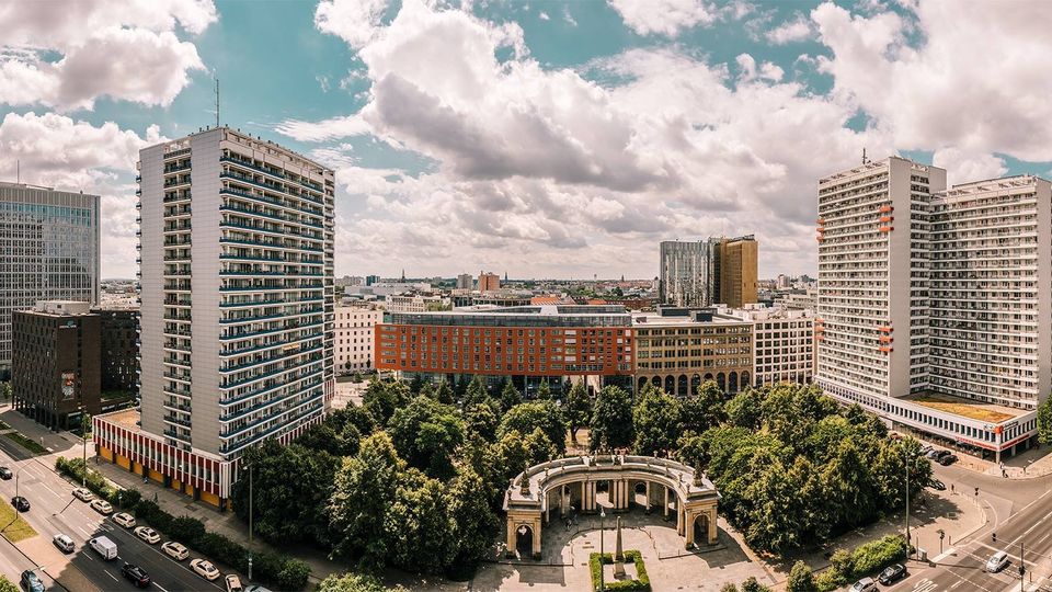 Panoramablick auf ein städtisches Viertel mit mehreren hohen Wohn- und Bürohochhäusern, einem langgestreckten Gebäuderiegel und breiten Straßen. Im Vordergrund liegt ein dicht bewachsener Park mit einem halbkreisförmigen, monumentalen Säulenbau und einem Platz.