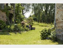 Ein grüner Garten mit Rasenfläche, umgeben von dichten Bäumen und blühenden Sträuchern. Im Vordergrund sind eine Ziegelwand und eine hölzerne Konstruktion zu sehen, die von Pflanzen bewachsen sind.