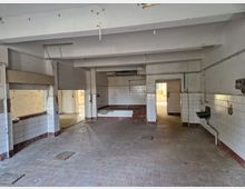 Leerer Innenraum eines alten Gebäudes, vermutlich eine ehemalige Küche oder Werkstatt, mit weißen, halbhohen Fliesen an den Wänden, einem gefliesten Boden und offenen Durchgängen. Einige installierte Waschbecken und sichtbare Rohrleitungen deuten auf einen früheren Gebrauch hin.