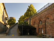 Backsteinbau mit Rundbogenfenstern und einem gepflasterten Weg, der von links nach oben führt, flankiert von einer Treppe und Geländern. Im Hintergrund ein runder Turm, umgeben von Bäumen, sowie eine überdachte Holzstruktur im Vordergrund.