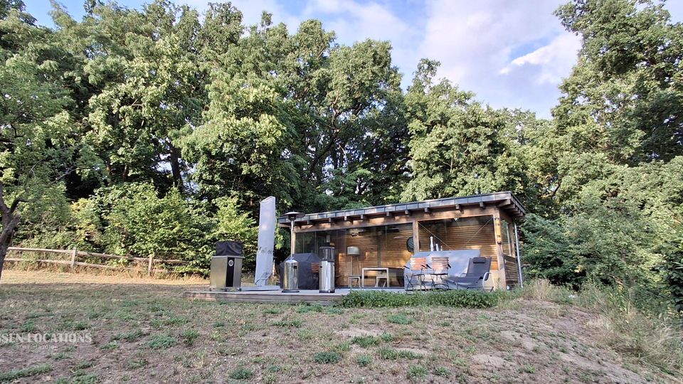 Kleines, modernes Holzhaus mit flachem Vordach und großer Glasfront auf einer Lichtung, davor eine Terrasse mit Außenmöbeln. Umgeben von dichtem Wald und Sträuchern, mit leicht abfallender Wiese im Vordergrund.