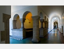 Langer Korridor mit Rundbögen auf massiven Steinsäulen, gelblich beleuchtetem Seiteneingang und einem dunkelgrünen Boden, umrahmt von weißen Wänden und einer gewölbten Decke. Die Beleuchtung erfolgt durch an der Decke hängende Lampen.