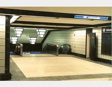 U-Bahn-Station mit gefliesten Wänden, einer Rolltreppe, die zu einem unteren Bahnsteig führt, und moderner Beleuchtung in geometrischer Form. Ein Hinweisschild mit der Aufschrift „Leinestraße“ ist über dem Treppenbereich angebracht.