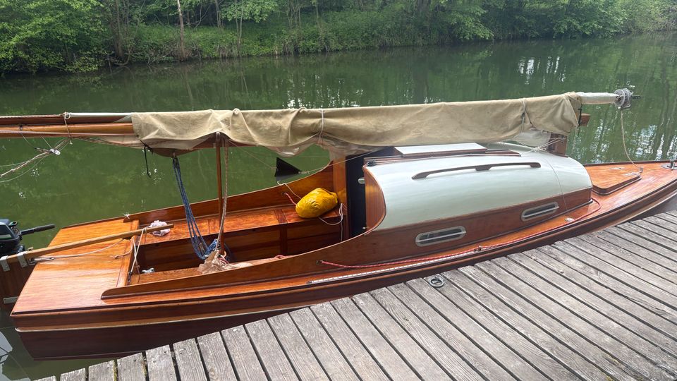 Holzsegelboot mit beige eingerolltem Segel liegt an einem Holzsteg auf einem ruhigen, grünlichen Fluss. Im Hintergrund säumen dicht bewachsene Ufer mit Bäumen und Sträuchern das Gewässer.