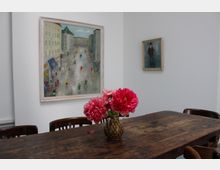 Ein heller Innenraum mit weißer Wand, an der zwei gerahmte Gemälde hängen. Im Vordergrund steht ein langer, dunkler Holztisch mit Stühlen und einer Vase mit pinken Blumen in der Mitte.