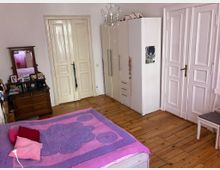 Schlafzimmer mit Holzboden, einem Bett mit rosa-violetter Bettdecke, einem weißen Kleiderschrank, einer Kommode aus Holz mit Spiegel und zwei weißen Doppeltüren. Eine kleine Lampe hängt von der Decke.