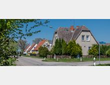 Straßenszene in einer Wohngegend mit Einfamilienhäusern, die Reihenhäuser mit Satteldächern und Zäunen zeigen. Die Umgebung ist geprägt von Gärten, Bäumen und einer asphaltierten Straße unter einem klaren blauen Himmel.