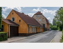 Straßenszene in einer Wohngegend mit zwei Gebäuden im Vordergrund; ein einstöckiges Haus mit orangener Fassade und braunen Ziegeldach und ein größerer Wohnbau im Hintergrund mit heller Putzfassade und roten Ziegeldach. Die Straße ist asphaltiert und von Gehwegen sowie Bäumen gesäumt.