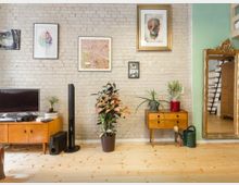 Wohnzimmer mit heller Ziegelwand, dekoriert mit mehreren Kunstwerken und Goldrahmen. Möbel im Mid-Century-Stil, darunter zwei Holzkommoden, Zimmerpflanzen, ein Fernseher und ein großer, goldener Standspiegel; daneben offener Zugang zu einer Treppe.