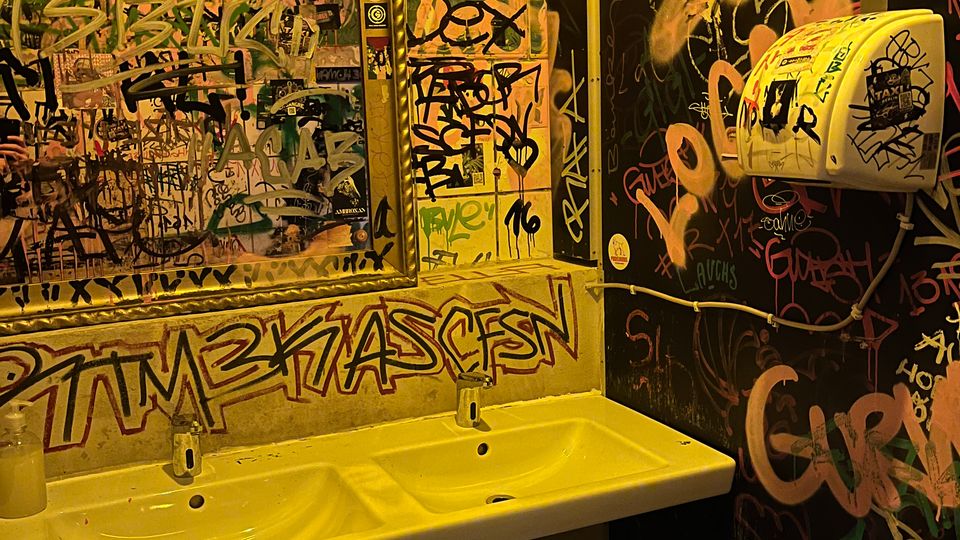 Kleiner Sanitärraum mit Doppelwaschbecken und großem Spiegel, dessen Rahmen und Glas mit zahlreichen Graffiti und Aufklebern bedeckt sind. Die Wände sind vollständig mit bunten Tags und Schriftzügen überzogen; rechts hängt ein Wandspender neben sichtbaren Leitungen.