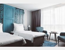 Helles Hotelzimmer mit zwei Einzelbetten, einem blauen, rustikal gestalteten Holzkopfteil, zwei blauen Polstersesseln, einem runden Beistelltisch und großen Fenstern mit lichtdurchlässigen Vorhängen. Der Boden ist mit Holzoptik und einem blauen Teppich gestaltet.