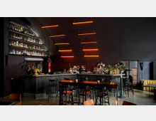 Eine moderne Bar mit dunkler Einrichtung, halbkreisförmigen Theken und schwarzen Barhockern. Im Hintergrund befinden sich beleuchtete Regale mit Flaschen, auffällige orangefarbene Leuchtstäbe an der Decke und vereinzelte Pflanzen als Dekoration.