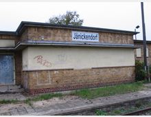Backsteinbahnhofsgebäude des Bahnhofs "Jänickendorf", mit graffitibeschmierten Wänden und einem leicht verwilderten Bahnsteig im Vordergrund. Links ist eine Tür zu sehen und rechts daneben ein beleuchteter Mast.