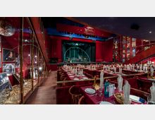 Ein Theater- und Veranstaltungsraum mit einer Bühne, roten Vorhängen und einem darüber hängenden Schild mit der Aufschrift "Wintergarten". Der Saal ist mit langen, festlich gedeckten Tischen bestückt, umgeben von roten Stuhlpolstern. An den Seitenwänden sind beleuchtete Plakate und Fotos ausgestellt.