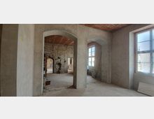 Ein unfertiger Innenraum mit Sicht auf rohe Steinwände, Rundbögen und unverputzte Flächen. Durch große, teilweise abgedeckte Fenster fällt Tageslicht, und im Raum befinden sich Bauwerkzeuge und Materialien.