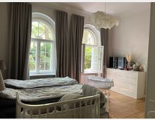 Ein Schlafzimmer mit großen, halbrunden Fenstern, die von langen Vorhängen eingerahmt sind. Im Raum stehen ein Doppelbett, ein Babybett und eine weiße Kommode mit Fernseher, auf Holzboden und mit Blick auf einen grünen Garten.