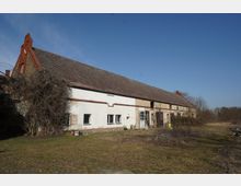 Langes, einstöckiges Wirtschaftsgebäude mit stark geneigtem Ziegeldach, teils verputzter und teils freigelegter Backsteinfassade sowie mehreren Fenstern und Toröffnungen. Davor liegt eine verwilderte Hoffläche mit Büschen und kahlen Bäumen am Rand einer ländlichen Umgebung unter klarem Himmel.