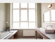 Helles Badezimmer mit großer Fensterfront, modernen weißen Fliesen an den Wänden und Waschbecken auf einer Arbeitsplatte aus Holz. Auf der linken Seite befindet sich eine eingelassene Badewanne und eine kleine Orchidee als Dekoration.