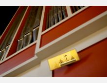 Nahaufnahme einer Kirchenorgel mit roten und weißen Umrahmungen sowie silbernen Orgelpfeifen. Im unteren Bereich der Aufnahme ist ein beleuchteter Ausschnitt eines Altars mit einem Kreuz und Blumenschmuck zu sehen.