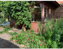 Ein kleiner Gartenbereich mit einem Blumenbeet voller bunter Blumen vor einem alten Gebäude aus rotem Backstein und Holz mit großen Fenstern. Links steht eine Metallbank auf einem gepflasterten Weg, umgeben von dichtem Grün.