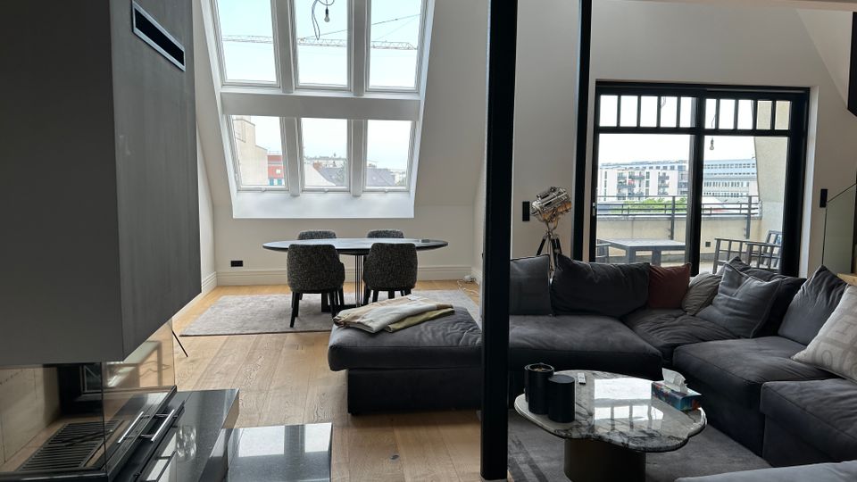 Modernes, offenes Wohnzimmer mit Essbereich unter einer Dachschräge, großen Fensterflächen und hellem Holzboden. Im Vordergrund stehen ein graues Ecksofa mit Marmorcouchtisch, daneben ein runder Esstisch; rechts führt eine Glastür auf einen Balkon.