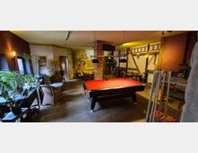 Innenraum eines rustikalen Aufenthalts- oder Gastraums mit rotem Billardtisch unter drei Hängelampen, Backsteinsäule und Holzbalken an den Wänden. Links stehen Pflanzen und Korbsessel an einer Fensterfront, rechts sind Tische, Stühle und ein Billardqueue-Ständer vor einer gemauerten Theke.