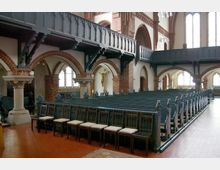 Innenansicht einer Kirche mit roten Ziegelbögen, hölzernen Kirchenbänken und einer umlaufenden schwarz gestrichenen Empore. Im Hintergrund ist ein Kruzifix sowie mehrere bunte Kirchenfenster zu sehen.