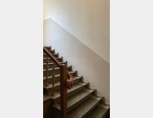 Treppenhaus mit abgenutzten Steinstufen, einem hölzernen Handlauf und schlicht gestalteten, hellen Wänden. Das Geländer besteht aus vertikalen Holzstreben.