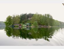 Eine grüne Uferlandschaft mit mehreren kleinen Häusern, darunter ein auffälliges grünes Haus, die am Rand eines ruhigen Sees mit spiegelglatter Wasseroberfläche liegen. Im Hintergrund dichter Wald, der sich im Wasser reflektiert.