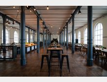 Geräumige Halle mit hohen Decken, aufgereihten Tischen und schwarzen Metallstühlen, umgeben von großen, gewölbten Fenstern. Der Raum wird von mehreren dunklen Säulen und hängenden Lichterketten geprägt.