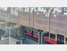 Außenansicht eines Bahnhofsbereichs mit rostfarbenen Metallverkleidungen, die mit Graffiti und Aufklebern bedeckt sind. Darunter fährt ein gelb-roter S-Bahn-Zug vorbei, während im Vordergrund Treppen, Betonwände und ein Geländer sichtbar sind.