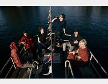 Eine Gruppe von sechs Personen sitzt und steht auf einem kleinen Boot auf einem Gewässer, umgeben von einer Reling. Bunte Wimpel sind auf einer Stange in der Mitte angebracht, und die Personen halten Flaschen in der Hand, während sie sich unterhalten.