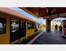 Ein gelber U-Bahn-Zug steht an einem überdachten Bahnhof mit gelbem Holzdach und einer großen Bahnhofsuhr. Im Hintergrund sind historische Gebäude sowie grüne Bäume zu sehen, und einige Menschen stehen am Bahnsteigende.