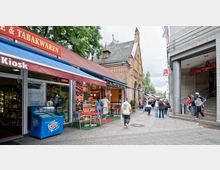Straßenszene mit einem Kiosk und einem Imbiss auf der linken Seite, einer historischen Backsteinfassade im Hintergrund und dem Eingang zu einem Einkaufszentrum ("Ring-Center") auf der rechten Seite. Mehrere Menschen sind auf dem Gehweg unterwegs oder stehen in Gruppen.