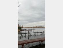 Ein See mit mehreren hölzernen Stegen im Vordergrund, umgeben von kahlen Bäumen und Sträuchern. Am Horizont ist ein kleiner Ort mit Gebäuden und mehreren Kirchtürmen zu sehen, unter einem bewölkten Himmel.