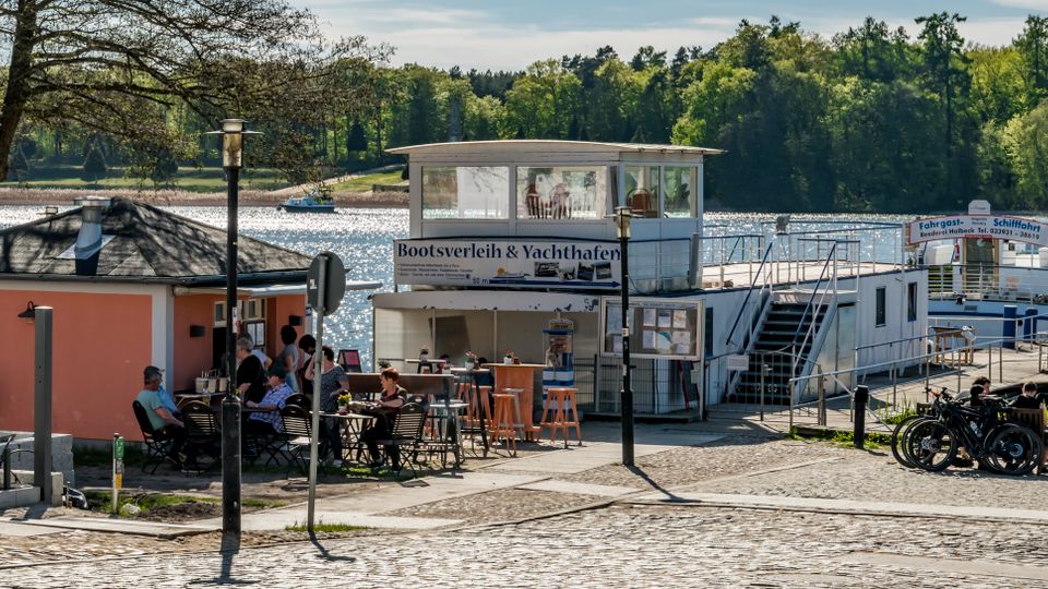 Lakeside marina area with a boat rental and yacht harbor building, outdoor seating, and a docked passenger boat beside a pier. Cobblestone waterfront promenade with railings, bicycles, and trees along the water with forested hills in the background.