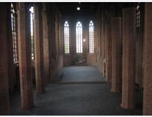 Innenansicht einer gotischen Backsteinkirche mit hohen, spitzbogigen Fenstern und massiven Säulen. Der Raum ist schlicht gehalten, mit einem dunklen Steinboden und einem kleinen Altarbereich im Hintergrund.