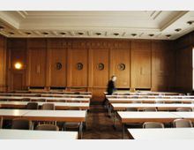Ein holzvertäfelter Seminarraum mit mehreren Reihen schlichter Tische und Stühle. Die Wand im Hintergrund ist mit runden Verzierungen und einem klassischen Türrahmen gestaltet, während die Decke mit eingelassenen Leuchten strukturiert ist.