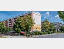 Wohngebiet mit mehrstöckigen Wohngebäuden, umgeben von Bäumen und Grünanlagen. Im Vordergrund sind ein kleiner Parkplatz mit geparkten Autos, ein Kopfsteinpflasterweg und ein niedriger Zaun erkennbar.